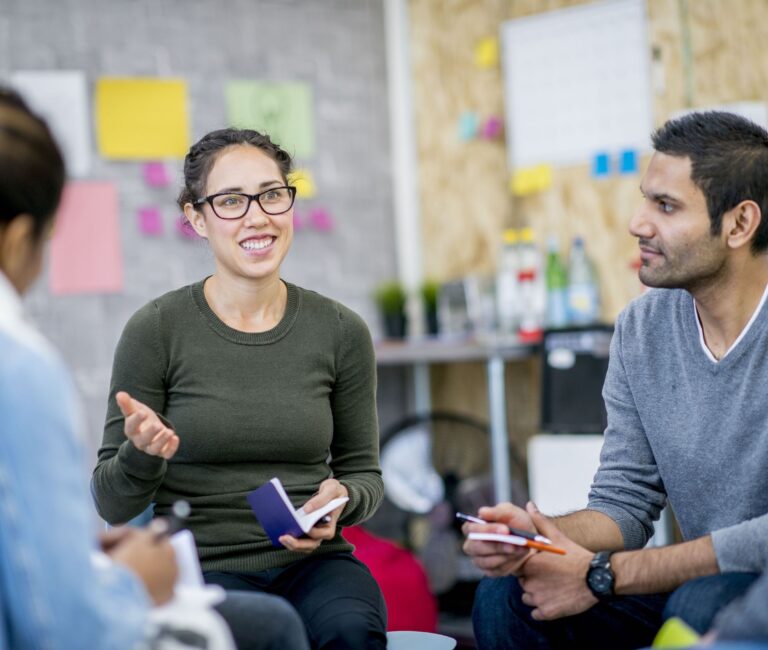 A group of entrepreneurs are indoors in a workshop. They are sitting and brainstorming new ideas. They are holding pens and notebooks to write down ideas. One woman is leading their group discussion
